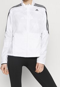 Chaqueta deportiva blanca hecha de una tela suave y liviana. Presenta un diseño de tres franjas negras en las mangas y bolsillos frontales con cremallera.