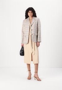 Blazer à carreaux beige, à boutonnage simple avec des boutons marron, sur une robe beige ceinturée. Accessoirisé avec un sac à main noir et des sandales à lanières beiges.