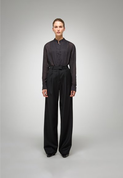 Zwarte high-waisted broek met scherpe plooien, gecombineerd met een donker, doorschijnend overhemd met knopen. Gladde, gestructureerde stoffen en strakke lijnen.