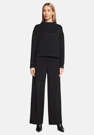 Woman with straight blonde hair wears black long-sleeve top with embellished text, wide-leg black pants, and black pointed boots, standing.