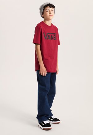 Red cotton T-shirt with black "VANS" logo, paired with blue denim pants and black sneakers with white accents and gum soles. Gray beanie.