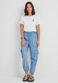 White crew-neck t-shirt with green logo, paired with light blue cargo pants featuring elastic cuffs and side pockets. Worn with metallic sandals.
