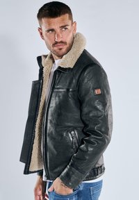 Schwarze Lederjacke mit einem hellbraunen Lammfellkragen, ausgestattet mit Reißverschlusstaschen und einer strukturierten Oberfläche, gestaltet mit einem taillierten Gürtel.