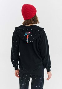 Czarna bluza z kapturem w białe kropki, ze sznurkiem i metką z tyłu. Połączona z dopasowanymi spodniami w czarne kropki oraz czerwoną czapką beanie.