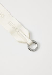White fabric strap with "HUGO" embossed, featuring a silver metal ring attachment. Smooth texture, minimalist design.