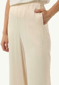 Beżowe spodnie o szerokich nogawkach z elastycznym paskiem i bocznymi kieszeniami. Gładki, lekki materiał o subtelny blasku.
