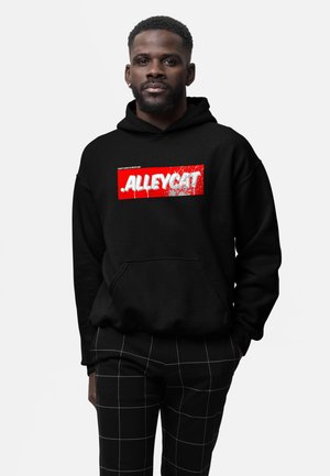 Mężczyzna ubrany w czarną bluzę z kapturem z czerwono-białym logo „ALLEYCAT” oraz czarne spodnie z białym wzorem w kratkę, stojący na jednolitym tle.