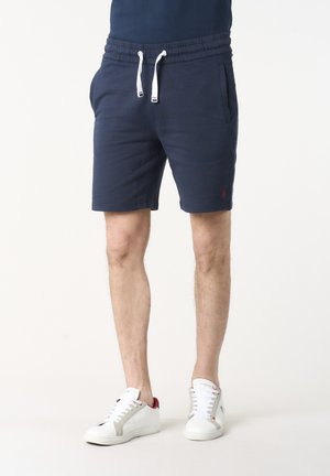 Uomo con pantaloncini blu navy con coulisse e sneakers bianche, in piedi davanti a uno sfondo bianco uniforme.