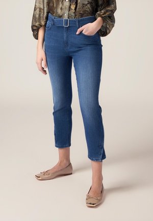 Blaue Denim-Capri-Jeans mit einem taillierten Bund, seitlichen Taschen und Knopfdetails am Saum. Kombiniert mit hellbeigen flachen Schuhen.