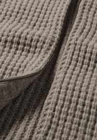 Grauer Strickstoff mit einem strukturierten Muster und einem silbernen Reißverschlussteil entlang einer Kante. Das Material wirkt weich und dick.