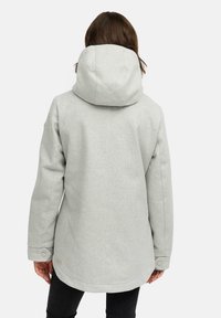 Femme portant une veste à capuche gris clair de dos, avec de longs cheveux bruns visibles, se tenant devant un fond uni.