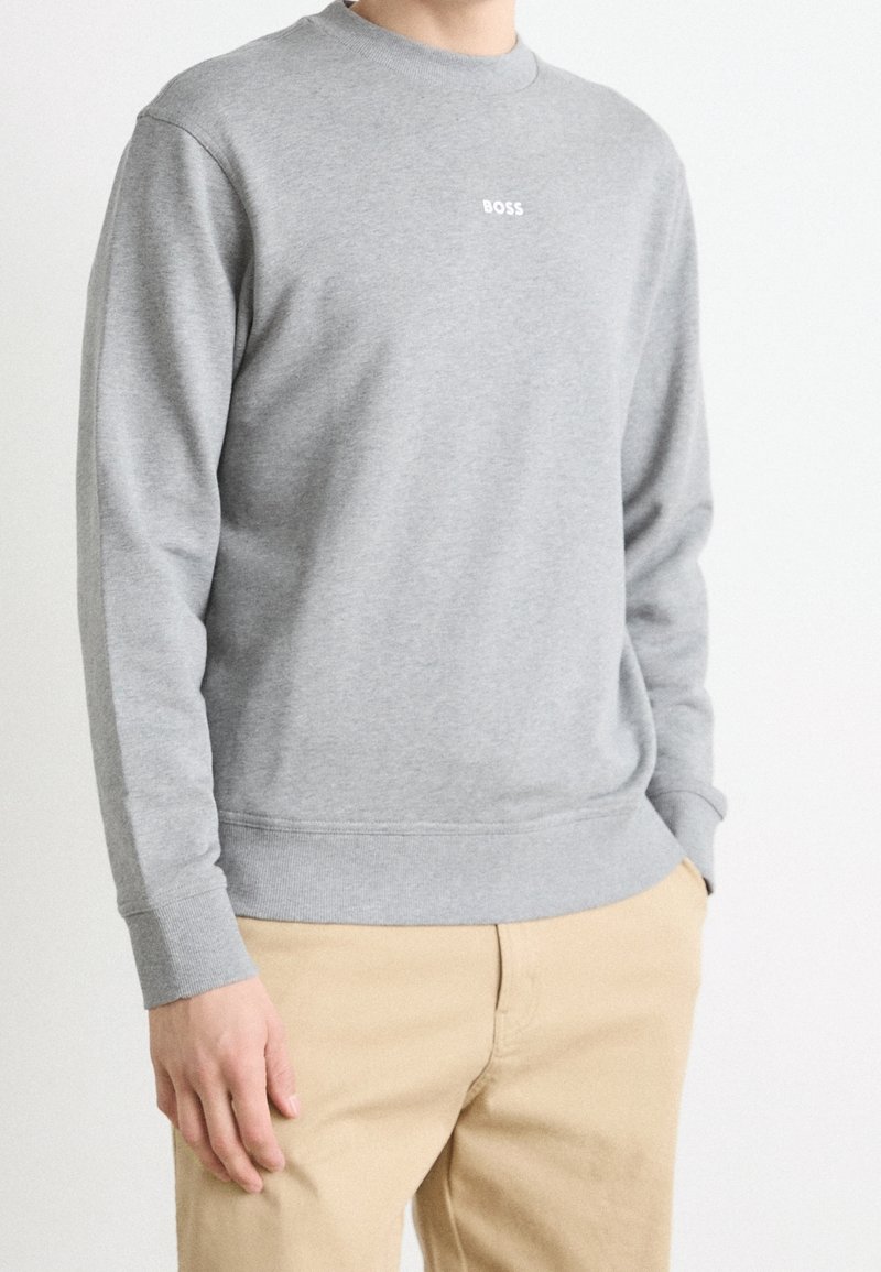 Hombre vestido con un sudadera gris claro con la palabra "BOSS" en blanco en el pecho y pantalones beiges, de pie con una mano en el bolsillo.