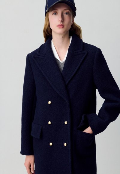 Cappotto doppiopetto navy in tessuto testurizzato, con bottoni dorati, ampie rever e tasche laterali. Indossato con un maglione grigio sotto.