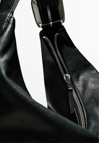 Schwarze Lederhandtasche mit glatter Textur, ausgestattet mit einem auffälligen silbernen Reißverschluss, metallischen Beschlägen und einer geschwungenen Form.