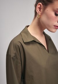 Chemise olive à coupe décontractée, avec un col pointu, un décolleté en V et des coutures vertes contrastantes le long des bords du col.