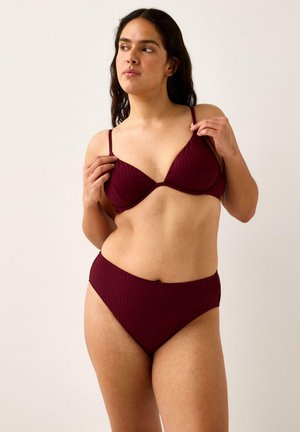 Kvinde, der justerer stropperne på en ribbet mørkerød bh og matchende høj talje-trusser foran en ensartet beige baggrund.