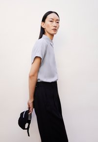 Lichtgrijs T-shirt met korte mouwen, gecombineerd met zwarte broek. De persoon houdt een marineblauwe pet met witte tekst vast en staat tegen een neutrale achtergrond.