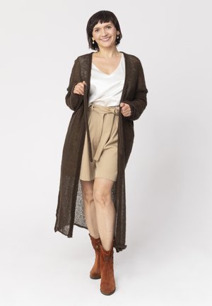 Kobieta uśmiechnięta, ubrana w długi brązowy kardigan, białą bluzkę, beżowe szorty z paskiem oraz brązowe zamszowe botki, stoi na białym tle.