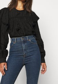 Blusa negra texturada con mangas acampanadas combinada con jeans de mezclilla azul de tiro alto. El conjunto presenta costuras prominentes y un frente de botones.