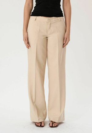 Žena stojaca pred jednoduchým pozadím, má na sebe béžové nohavice s širokými nohavicami, čierny tielkový top a hnedé sandále s otvorenou špičkou.