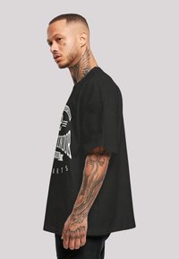 Mustad must shirt, millel valge graafiline trükk. Lühikesed varrukad, ümar kaelus, valmistatud puuvillast. Kätel on nähtav tätoveering.