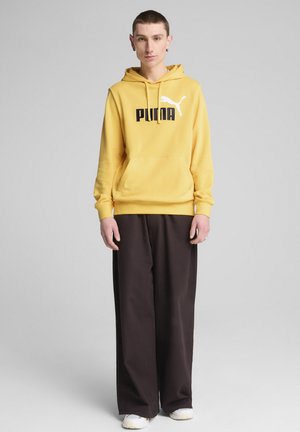 Sudadera amarilla con el logo negro de "PUMA" y una silueta blanca, combinada con pantalones holgados de color marrón oscuro y zapatillas blancas. Diseño minimalista.