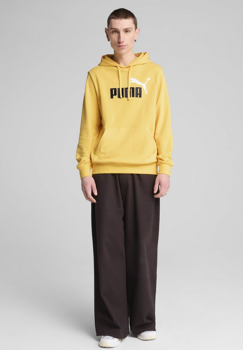 Felpa gialla con logo "PUMA" nero e silhouette bianca, abbinata a pantaloni larghi marroni scuri e sneaker bianche. Design minimalista.