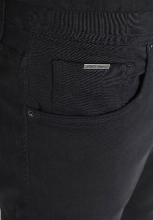 Close-up van zwarte spijkerbroeken met gedetailleerde stiksels, een klinknagel en een klein zakje met een metalen merketiket.