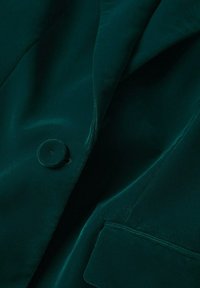 Veston en velours vert foncé avec un bouton recouvert de tissu, un revers et un détail de poche cousu.