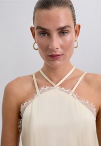 Camisole de couleur crème avec une bordure en dentelle au niveau du col. Présente des fines bretelles et une texture lisse et satinée, créant une apparence délicate.