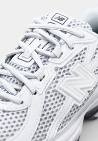 Vit sportig sko med meshupp. Vadderad tunga och gummisula. Har texturerade accenter och en framträdande logotyp på sidan.