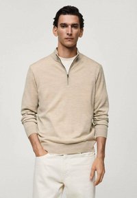 Beige zip-up sweater made of soft material, featuring long sleeves, a high collar, and ribbed cuffs, paired with white trousers.