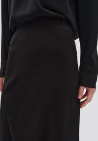 Personne portant un haut noir à manches longues et une jupe noire taille haute avec une couture verticale visible, la main reposant le long du côté de la jupe.