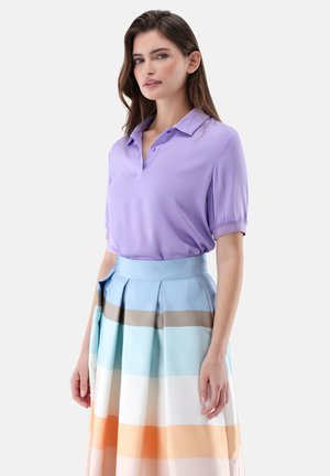 Frau trägt eine kurzärmelige lavendelfarbene Bluse, die in einen hoch taillierten, plissierten Rock mit horizontalen Pastellstreifen gesteckt ist.