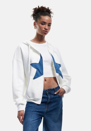 Witte rits hoodie met blauwe sterapplicatie, gecombineerd met een witte cropped top en high-waisted blauwe spijkerbroek. Gladde stoftextuur.