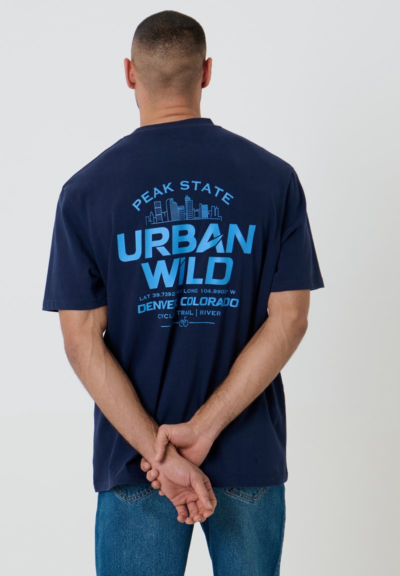 Hombre con camiseta azul marino con texto azul "Peak State Urban Wild Denver Colorado" y un gráfico del skyline de la ciudad en la parte trasera, con las manos entrelazadas detrás.