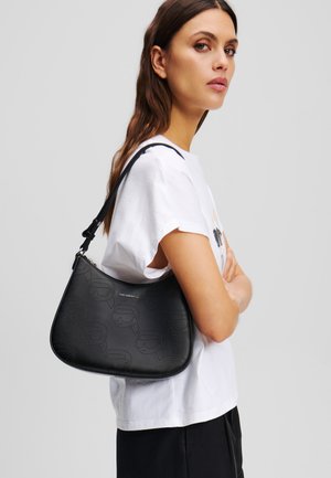 Sac à main en cuir noir avec un motif en relief, forme courbée, bandoulière réglable et fermeture éclair, porté sur l'épaule.