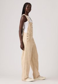 Beige denim overalls with wide legs, front pocket, and metal button accents, paired with a white tank top and white sneakers.
