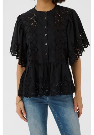 Mujer con blusa negra de manga corta con cortes de encaje y botones, combinada con vaqueros denim azul claro y una pulsera dorada.