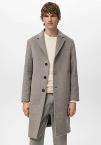 Grijze visgraatjas met reverskraag, drie zwarte knopen, zijzakken en een rechte snit. Draagt over een lichte trui en jeans.