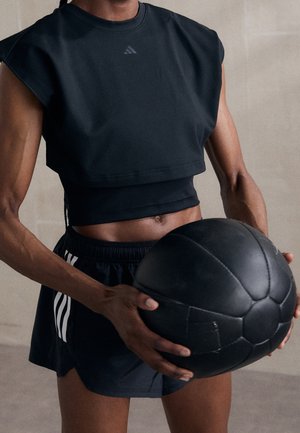 Sportovec v černém sportovním oblečení drží velký černý medicinbal v úrovni pasu na neutrálním pozadí.