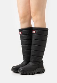 Svarta isolerade stövlar med en puffig design, försedd med texturerade paneler och en kraftig gummisula. Logotyper i röd och vit på toppen.