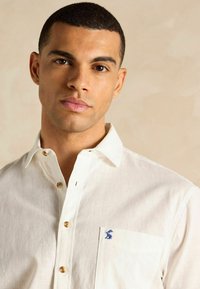 Balta linu krekls ar pogu apkakli, ar koka pogām un mazu zilu izsūktus logotipu uz krūšu kabatas.