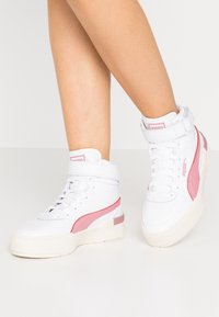 White high-top sneakers with pink accents, textured leather, strap around the ankle, and a thick cream rubber sole.
