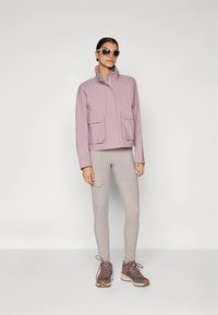 Veste violette claire avec deux poches avant, associée à un pantalon beige clair et des chaussures de sport marron. La veste est dotée d'un col montant.