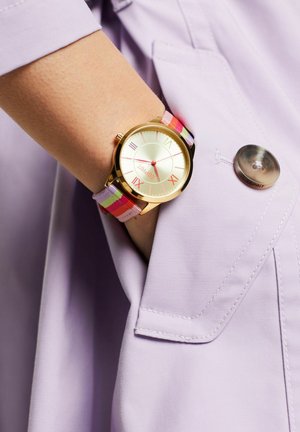 Goldene Uhr mit rundem Zifferblatt, cremefarbener Ziffernblatt, farbigen Streifenstoffarmband, auf einem lila Ärmel positioniert, mit einem silbernen Knopfdetail.