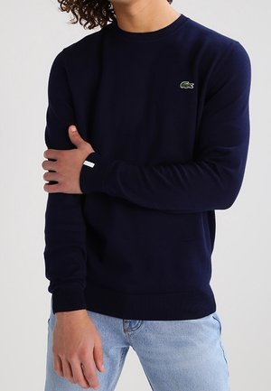 Mężczyzna ubrany w granatowy sweter z długim rękawem z małym zielonym logo krokodyla na piersi oraz jasnoniebieskie dżinsy.