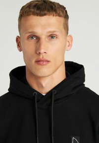 Sudadera negra con cordones, que presenta un pequeño logo blanco en un parche. Tejido suave, corte casual, cuello alto y diseño minimalista.