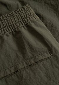 Gros plan sur un tissu vert olive avec une taille élastique et une poche arrière rectangulaire cousue sur un matériau texturé.