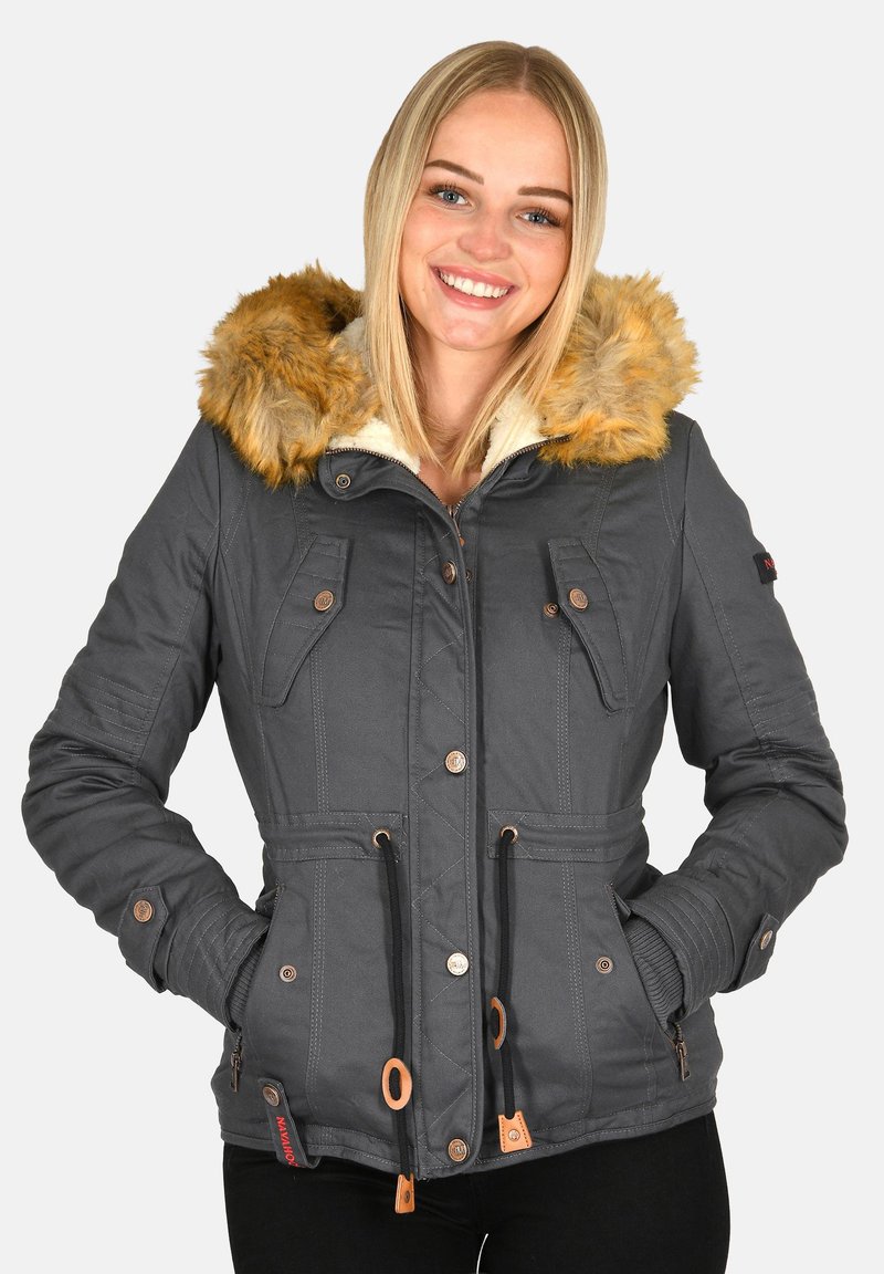 Smiling woman wearing a dark gray winter jacket with fur-lined hood, hands in pockets, and black pants against a plain background.